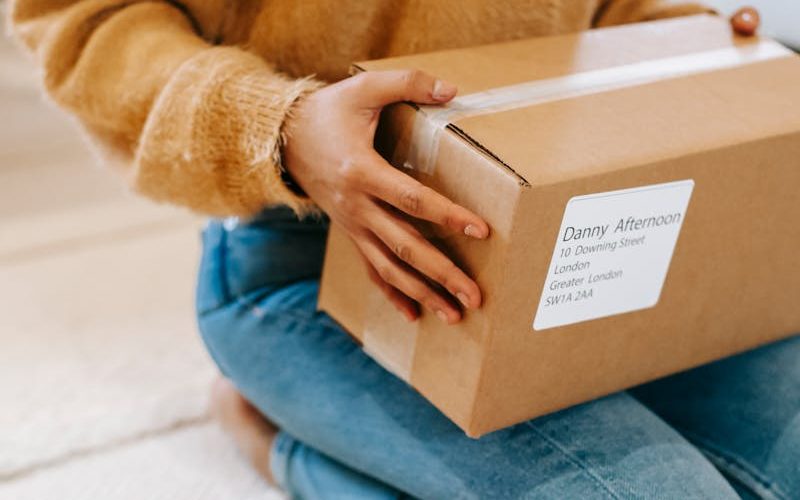Crop anonymous female in casual outfit sitting on floor with carton parcel on laps in light room